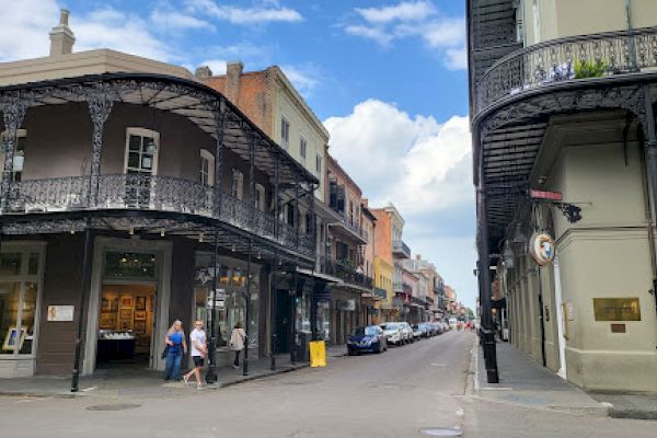 A street scene with historic buildings, featuring ironwork balconies, people walking, and cars parked along the road under a partly cloudy sky.