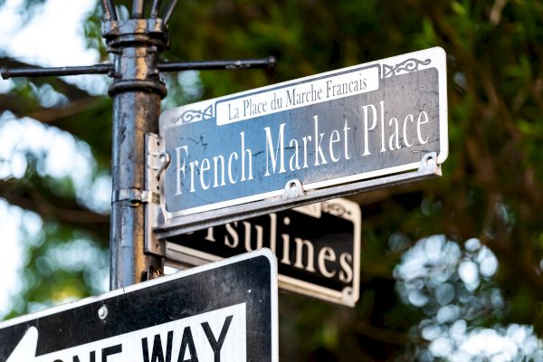 Street signs reading "French Market Place" and "Ursulines" with a "One Way" sign beneath, set against a leafy background.