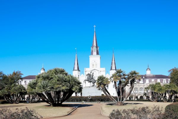 The image shows a park with a statue in the foreground and a cathedral with three spires in the background under a clear blue sky.