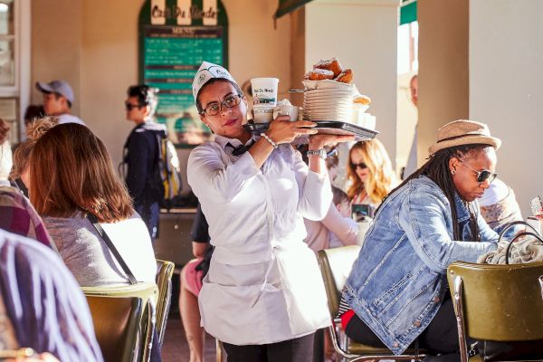 A waiter carries a tray through a busy outdoor cafe, with people seated at tables enjoying their meals and drinks.