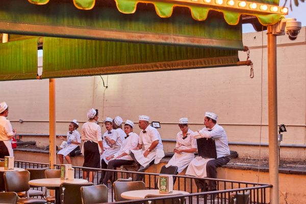 An outdoor seating area of Café Du Monde with several people in white uniforms sitting under an awning, and empty tables and chairs.