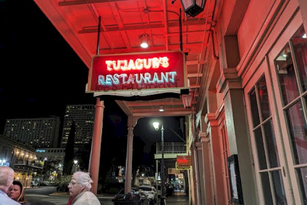 The image shows a street view with people and a lit sign reading "Tujague's Restaurant" under red neon lights at night.