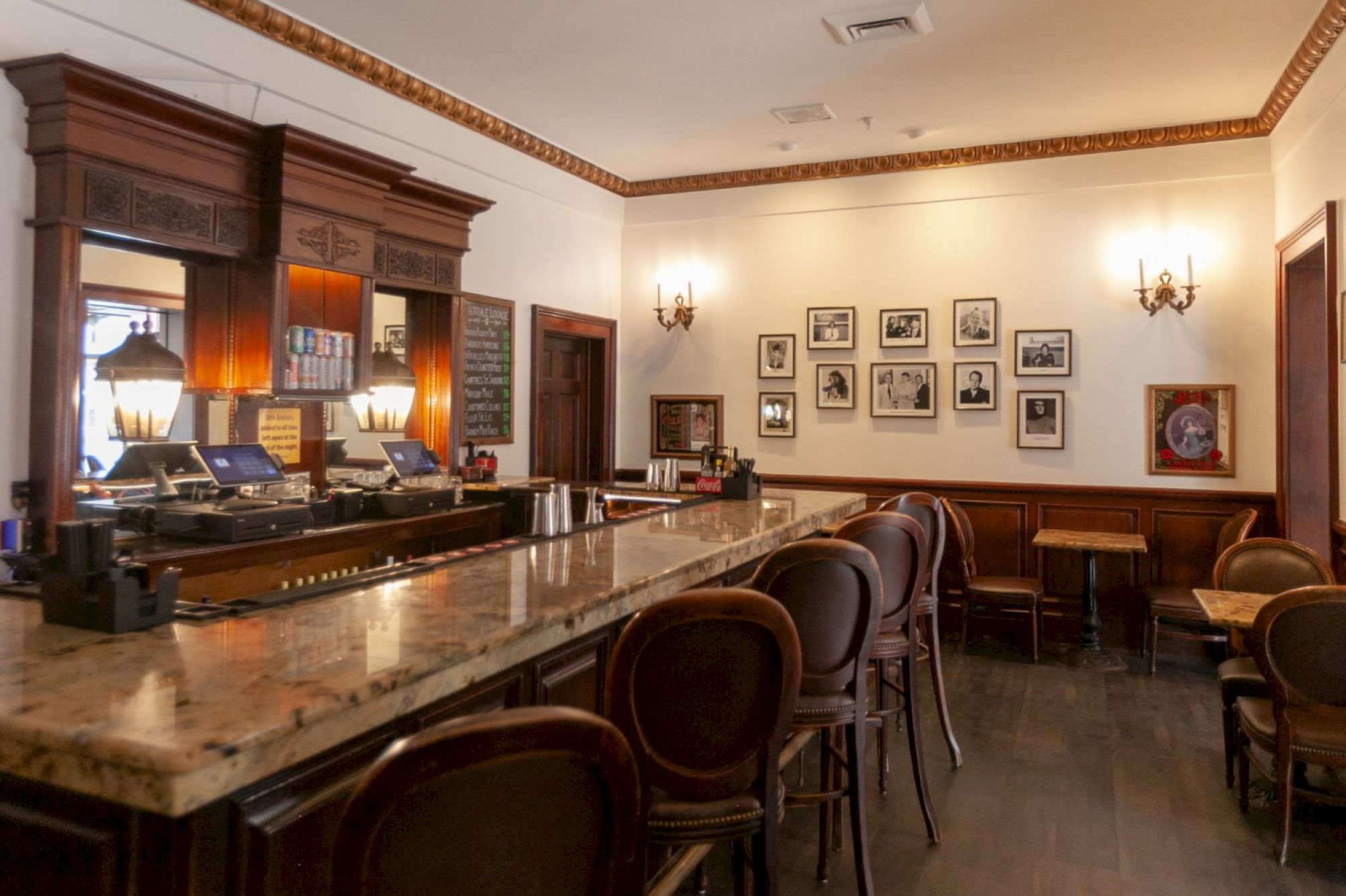 The image shows an empty bar with a marble counter, classic wooden chairs, and framed photos on the wall in a cozy setting.