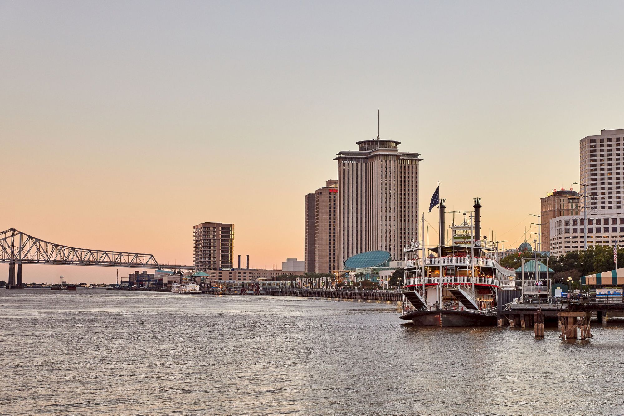 The image shows a riverside cityscape with buildings, a docked riverboat, and a bridge in the background during sunset.