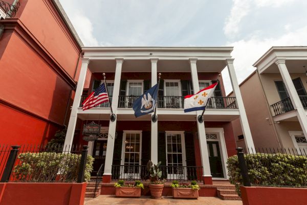 A two-story building with three flags displayed, featuring white columns, red walls, and a gated entrance with potted plants.