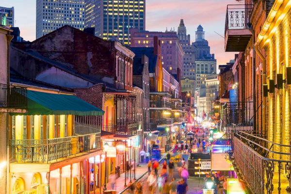 A bustling city street scene with colorful lights and classic architecture, possibly New Orleans, at dusk with people walking around.