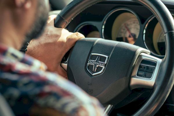 A person is driving a car, holding the steering wheel with a Dodge logo, wearing a patterned shirt.