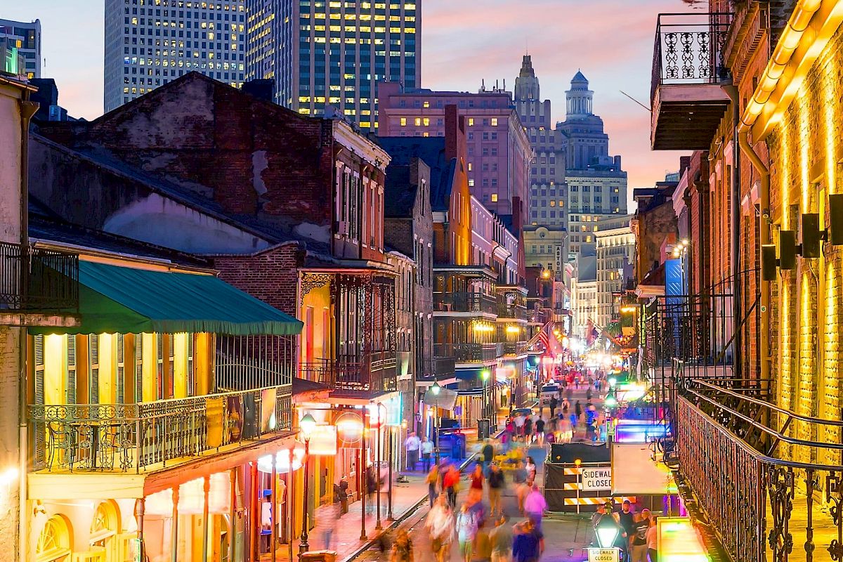 The image shows a lively street scene with colorful lights and blurred crowds, set against a backdrop of tall buildings at twilight.