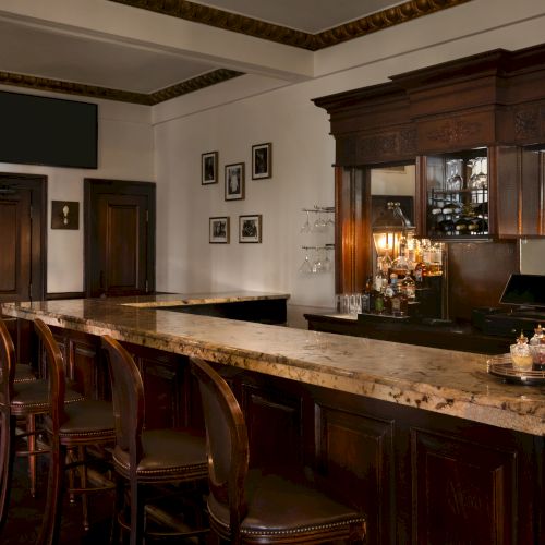 The image shows an elegant bar with wooden stools, marble countertops, framed pictures, and warm lighting, creating a classic atmosphere.