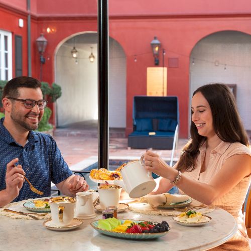 Two people are enjoying a meal at a round table by a pool, in a courtyard setting.