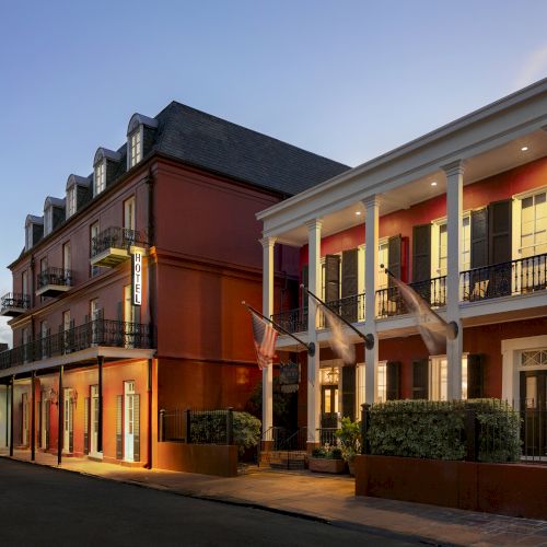 The image shows a two-story brick building with balconies and large columns, dimly lit at dusk, in a quiet street setting.