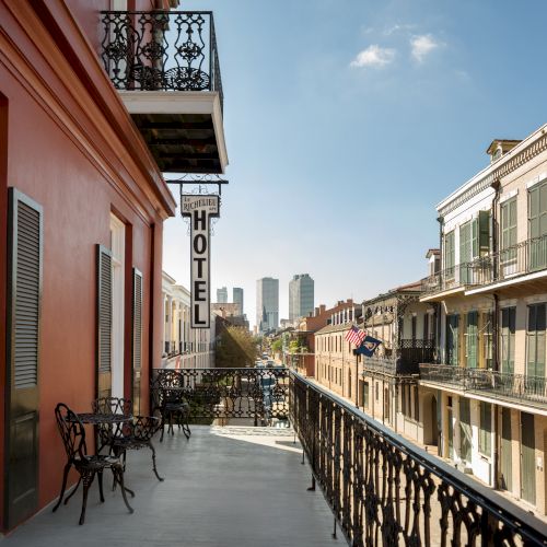 A balcony overlooks a street with historic buildings, featuring a "HOTEL" sign. The skyline is visible under a clear blue sky.