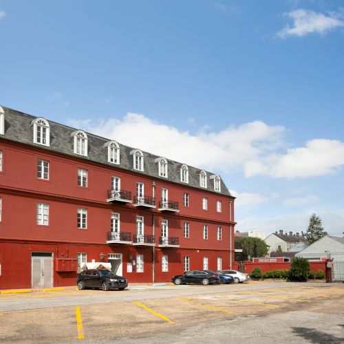 A red three-story building with cars parked in front, set against a clear blue sky with some clouds.