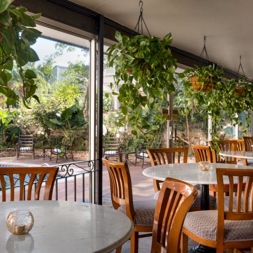 A cozy cafe with wooden chairs and marble tables overlooks a garden and pool, with hanging plants adding a touch of greenery.