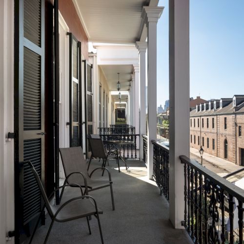 A balcony with chairs overlooks a street with brick buildings. The scene is under a clear blue sky, creating a peaceful atmosphere.