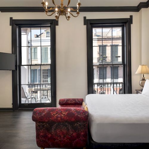 A hotel room with a large bed, red patterned bench, TV, dresser, lamps, and two tall windows displaying a view of a street outside.