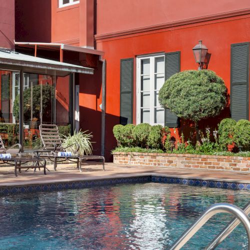 A red building with green shutters, a garden, and poolside loungers adjacent to a pool with metal handrails in the foreground.