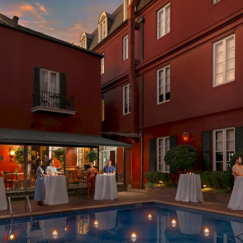 People are gathered around tall tables near a pool, with a red building in the background at dusk.