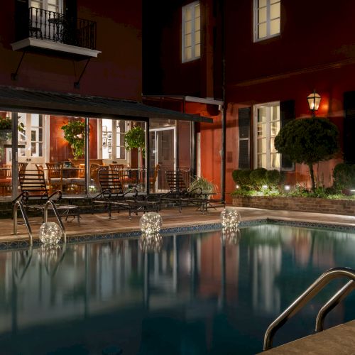 A nighttime view of a dimly lit outdoor pool area with chairs, tables, and a building with large windows in the background.