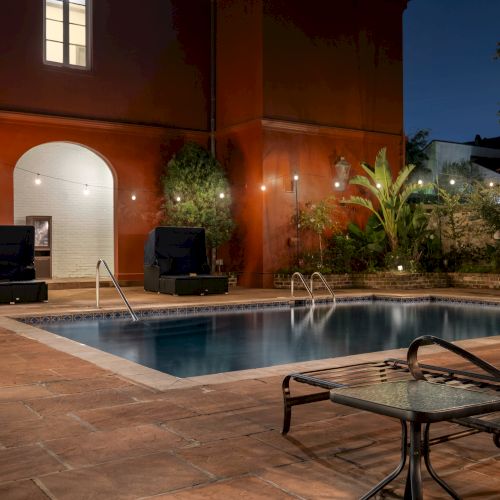 A nighttime scene showing a pool with lounge chairs, string lights, and a red building, surrounded by greenery.