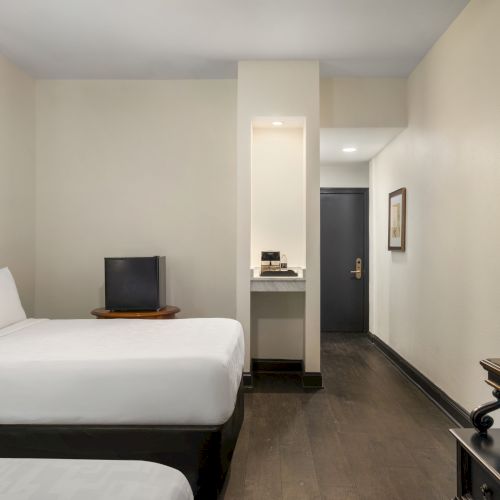 A hotel room with two beds, a wall-mounted TV, a small table, and a door in the background.