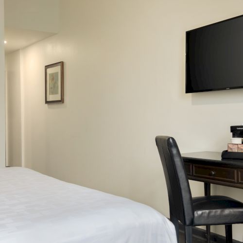 A hotel room with a bed, wall-mounted TV, framed art, desk, and chair with complimentary water and coffee setup on the desk.