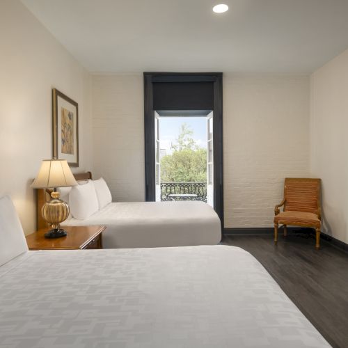 This image shows a hotel room with two beds, a lamp, a chair, a desk with a TV, and a window with a view.