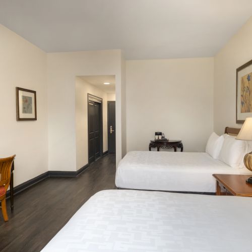 A hotel room with two beds, a lamp, TV, desk, chair, and artwork, featuring neutral tones and dark wood flooring.