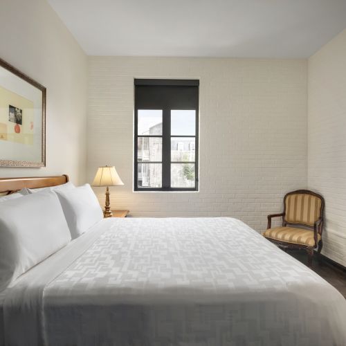 A neatly arranged bedroom with a bed, framed art, lamp, window, chair, and TV on the wall, all in a minimalistic style.