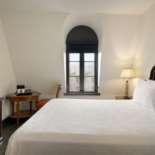 A hotel room with a large bed, a window, a side table with a lamp, a TV, a wooden desk, and a chair.