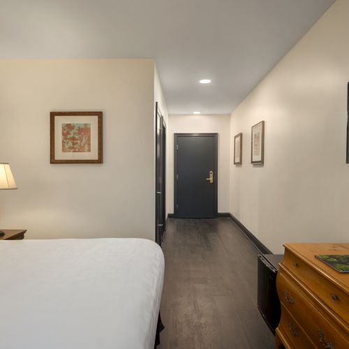 The image shows a hotel room with a bed, lamp, dresser, wall art, and TV, featuring wooden flooring and a closed door.