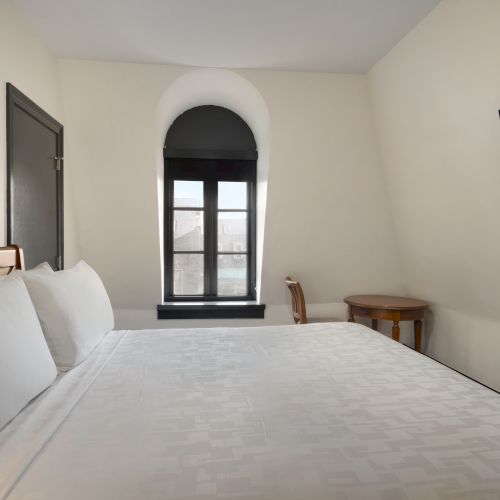 A neatly made bed in a room with a window, TV, and a small table with a chair against a plain wall.