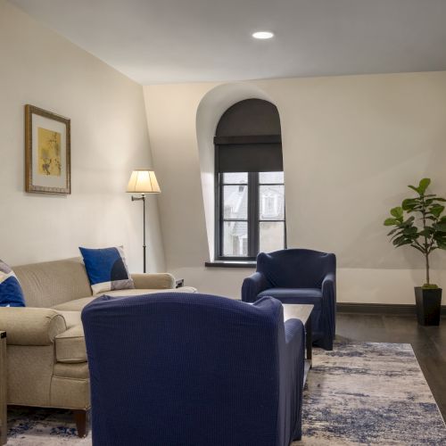 A cozy living room with blue chairs, a beige sofa, lamps, a potted plant, a window, and a wall-mounted TV, creating a relaxing ambiance.