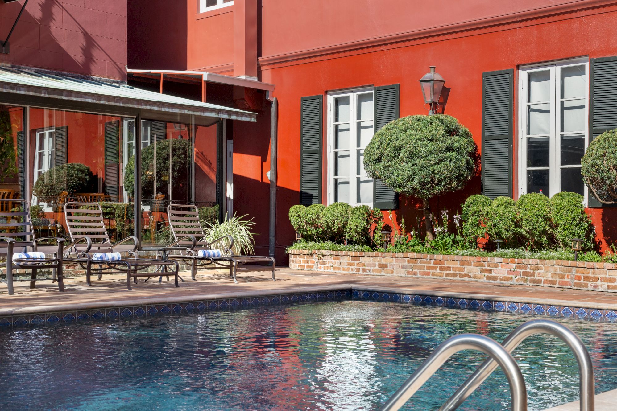 Outdoor pool with metal ladder, surrounded by lounge chairs, red building, and trimmed bushes. Sunny weather reflecting on the water.