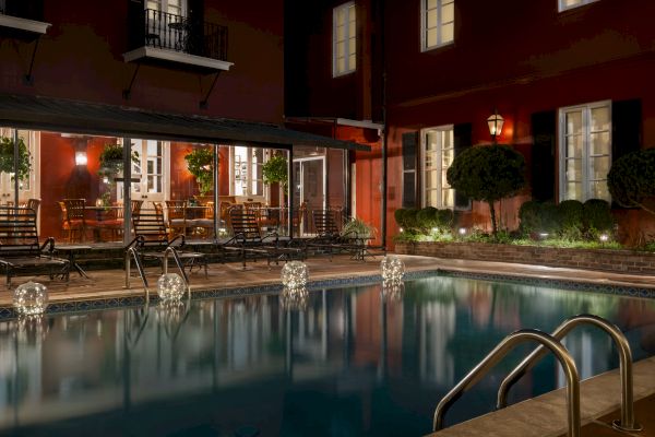 A dimly lit pool area with loungers, illuminated by globe lights, next to a plant-decorated patio with tables and chairs against a red building.